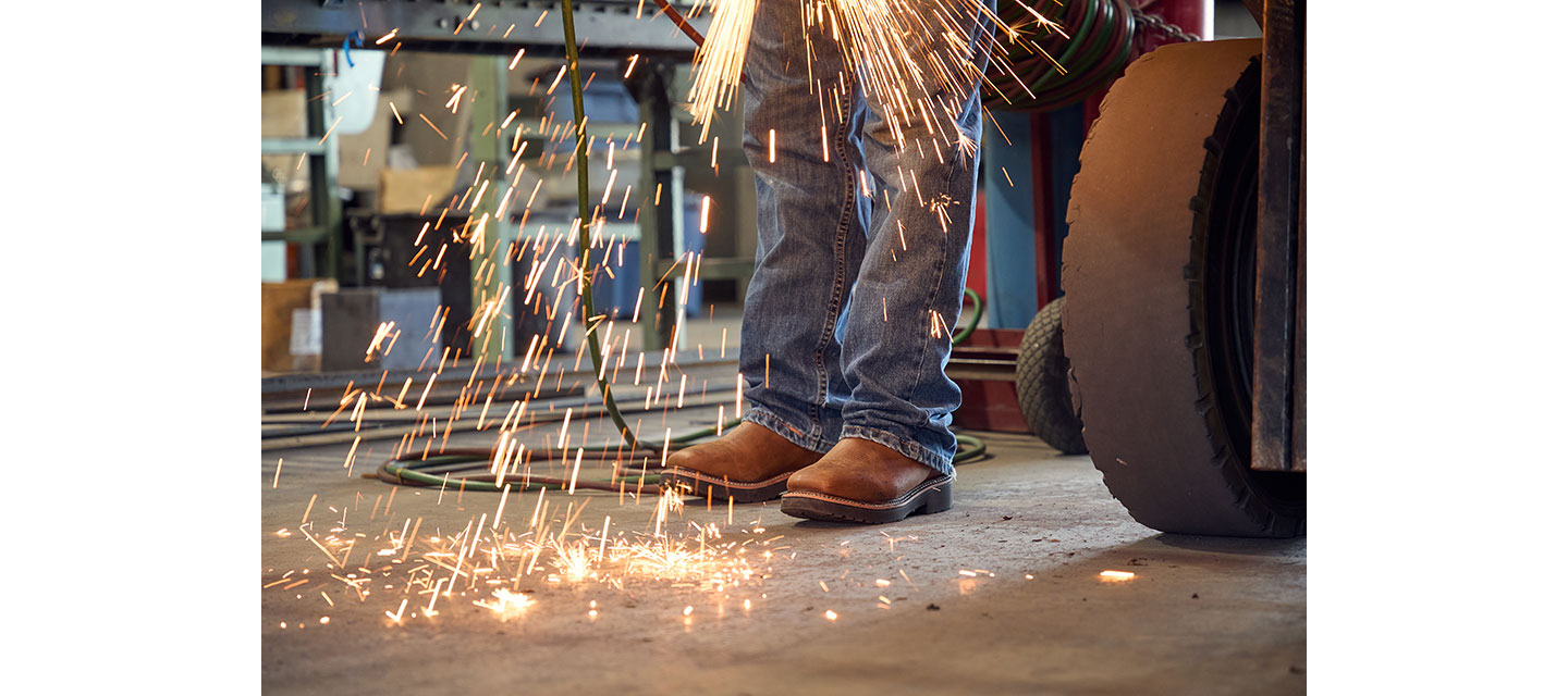 Las chispas saltan cuando Wes Stevenson, en jeans y botas de trabajo de cuero marrón, opera una herramienta de corte en un taller de metal, con el enfoque en sus piernas y pies.